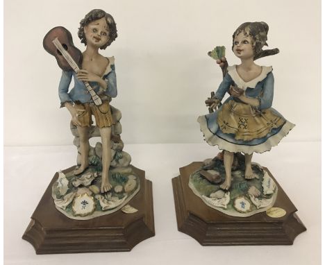 A pair of ceramic Capodimonte Bellinaso figurines of children, mounted onto wooden plinths.  A boy musician and a girl mendin