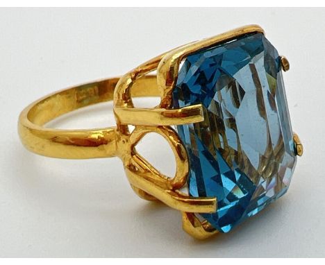 An 18k gold cocktail ring set with a large emerald cut Swiss blue topaz stone. Claw setting with decorative mount. Inside of 