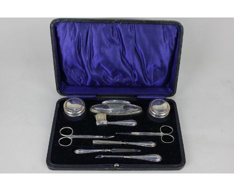 A George V cased silver manicure set in fitted case including a pair of silver topped glass jars, nail buffer, scissors and t