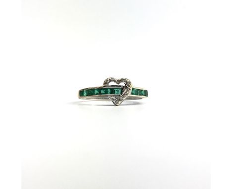 An 18ct white gold ring, set with a half hoop line of graduated calibre cut emeralds which passes through a diamond set heart