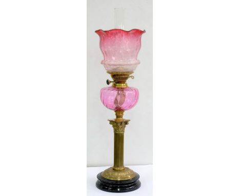 AN EDWARDIAN BRASS CORINTHIAN COLUMN OIL LAMP WITH EMBOSSED ROUND FOOT, CRANBERRY GLASS FOUNT AND BRASS BURNER, WITH A CRANBE