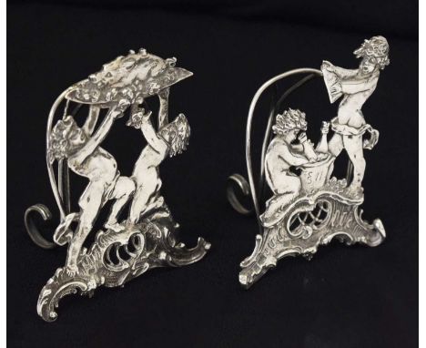 Pair German silver menu holders, one depicting cherubs with champagne bottles, the other depicting cherubs holding aloft a su