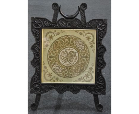 A late Victorian dark oak easel firescreen in the Aesthetic taste, with a needlework panel within a foliate carved frame.