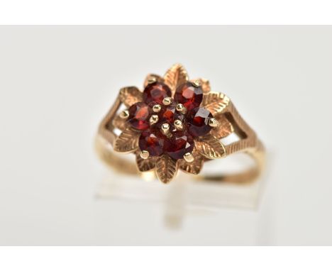 A 9CT GOLD GARNET DRESS RING, of a flower shape, set with seven circular cut garnets, textured leaf detailed surround, bifurc