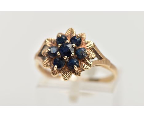 A 9CT GOLD SAPPHIRE CLUSTER DRESS RING, of a flower shape, set with seven circular cut blue sapphires, within textured leaf s