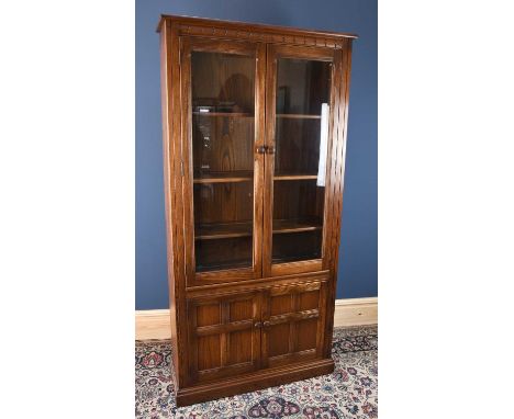 An Ercol style oak display cabinet with two glazed doors enclosing three adjustable shelves, above two panelled doors, on pli