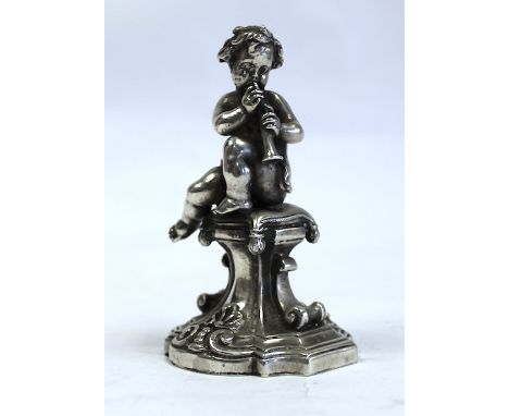  A Continental Silver Putto Figure depicted playing the flute seated on a cushion raised on a pedestal decorated with C-scrol