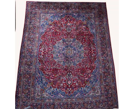 A BURGUNDY GROUND PERSIAN MASHAD CARPET, with centre floral blue medallion, on a burgundy ground with all-over pattern and li