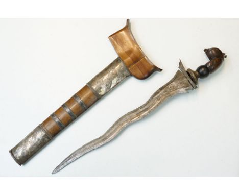 An Indonesian Kriss knife with traditional wave blade, carved wooden handle with wooden scabbard with white metal mounts.
