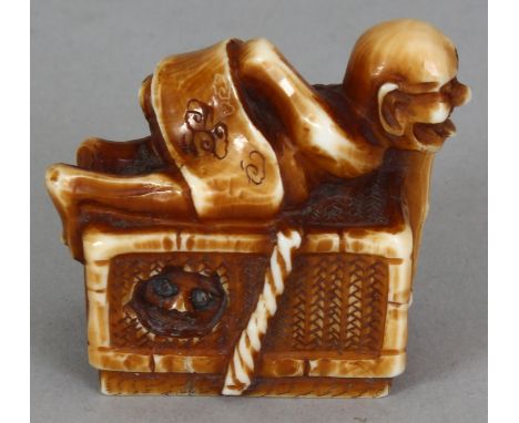 A SIGNED JAPANESE MEIJI PERIOD STAINED IVORY NETSUKE OF A DEMON, lying astride a rectangular reed woven box from out of which