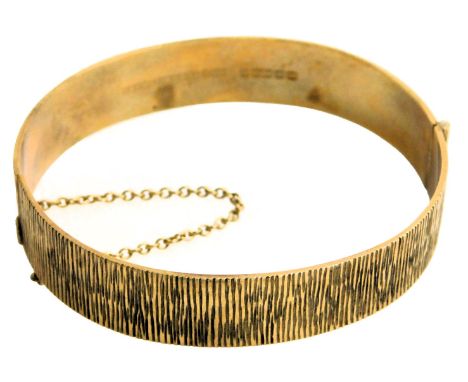A 9ct gold bangle, with a textured finish, on a snap clasp, with safety chain as fitted, 24.9g. 