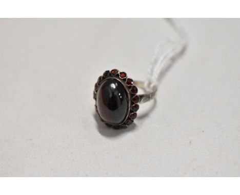 An oval garnet cabochon ring with garnet chip surround in a white metal collared mount on white metal loop, no marks, tests a
