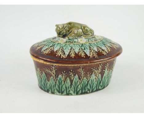A late 19thC Majolica potted meat dish and cover, with recumbent cow finial, leaf and floral moulding against a brown ground,