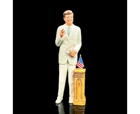 JFK in a crisp white suit with light blue tie holding books, standing next to a podium with a United States flag on top. Roya