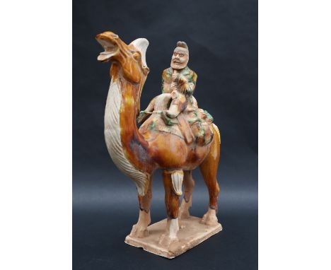 A Tang Dynasty style pottery statuette of a camel mounted with a bearded musician playing a shawm-like wind instrument, in br