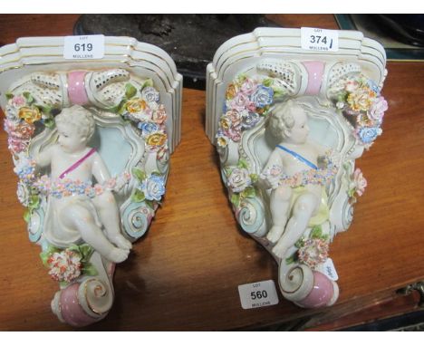 A PAIR OF PORCELAIN FLOWER ENCRUSTED WALL BRACKETS each with rectangular stepped shelf above a putti shown seated holding a g