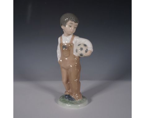 This NAO by Lladro figurine, titled Wanna Play? (1068), features a young boy dressed in overalls, holding a soccer ball under
