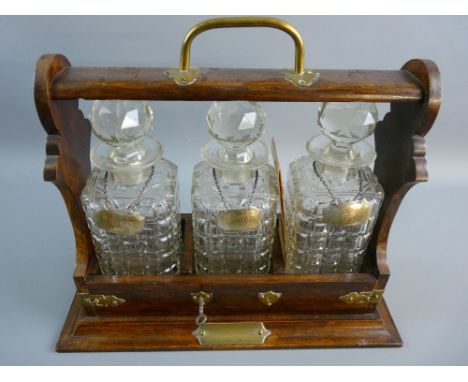 A VINTAGE OAK THREE BOTTLE TANTALUS with hallmarked silver labels having a brass carry handle and further embellishments, fro