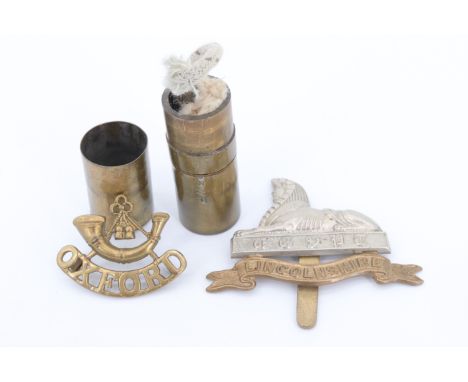 A trench art brass lipstick lighter, fabricated from a brass capacitor case, together with two cap badges, Oxford and Lincoln