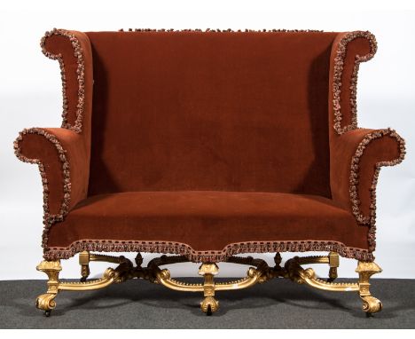 A George II style wing back sofa, upholstered in trimmed rust coloured velvet, gilt gesso under section with double incurved 