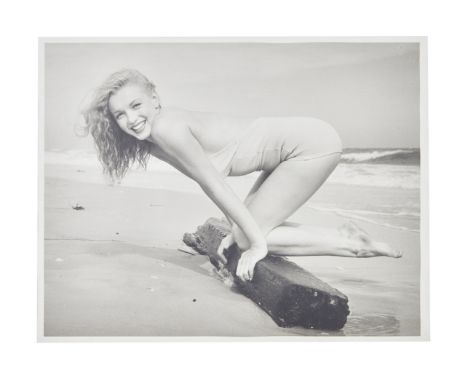 A black and white photograph of Marilyn Monroe wearing a swimsuit kneeling on a beach in Tobay Beach (Tobay being an acronym 