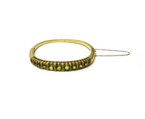 A 9ct gold, peridot and diamond set oval hinged bangle, the front mounted with a row of eleven circular cut peridots, graduat