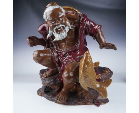 A striking porcelain or ceramic statue depicting an elderly fisherman crouching over a large golden carp. The fisherman, with