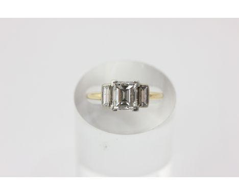 A diamond ring the rectangular step cut stone claw set with a rubover set baguette to each shoulder, the centre stone approxi