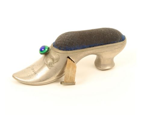 A white metal novelty tape measure in the form of a lady's shoe, blue velvet inset worn, the bow with glass boss and engraved