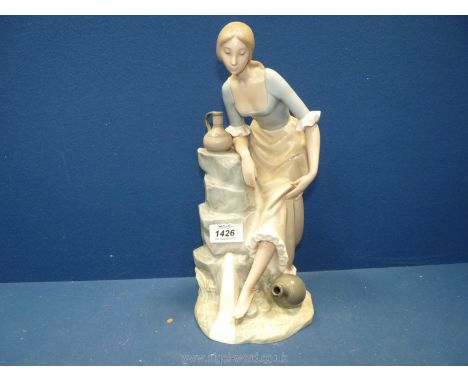 A Nao figure of a young girl holding her skirt and looking at her foot, with water vessels to her side, 13'' tall.