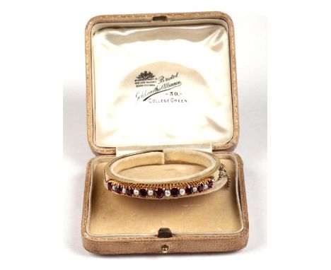A Victorian yellow metal ruby and pearl bangle of hinged circular form, 6cms diameter.