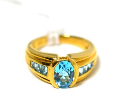 A topaz ring, an oval cut blue topaz in a yellow half rubbed over setting, to channel set step cut blue topaz shoulders, on a