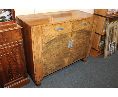 An Art Deco walnut sideboard, with rectangular top, and two drawers above two panel doors enclosing cupboard, with chrome han