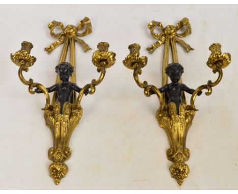 A pair of gilt metal and painted cherubic two branch wall sconces, the ribbon tied pediment above cherubic figures holding th
