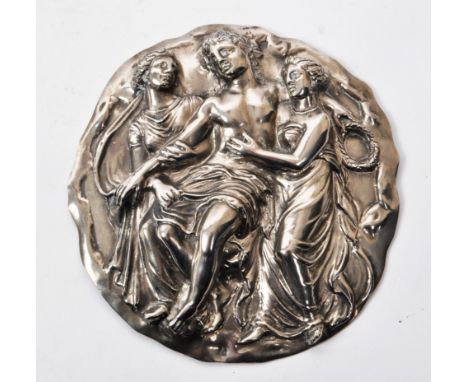 A vintage 20th-century white metal circular plaque, depicting a classical scene of a man in the middle of two women embracing