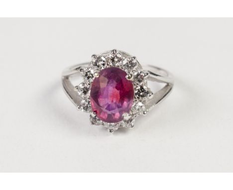 RUBY AND DIAMOND OVAL CLUSTER RING, with centre oval ruby and surround of twelve round brilliant cut diamonds, the white meta