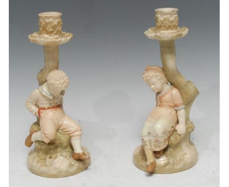 A pair of Royal Worcester figural candlesticks,  modelled by James Hadley, as a boy and girl sitting upon a rock beneath a tr