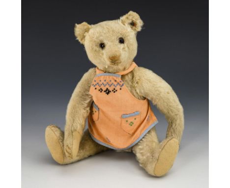 Alter Steiff-Teddy mit Schürze. Knopf im Ohr, mit rotem Fähnchenrest. Um 1920.Mohairplüsch. L 32 cm. Weißblonder Bär mit hell