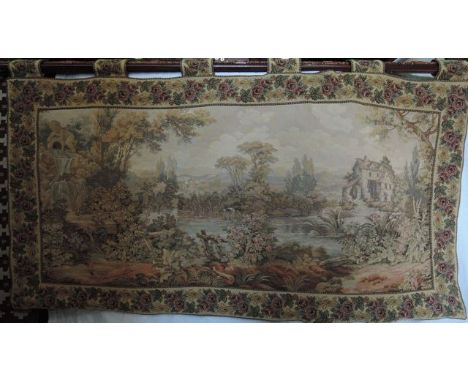 A machine woven tapestry wall hanging with river landscape, water mill, cherub and further detail within a floral running bor
