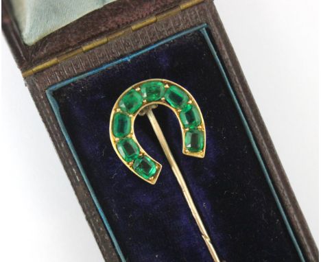 An early 20th century untested emerald and green stone stick pin, the horse shoe shaped head set with eight cushion cut green