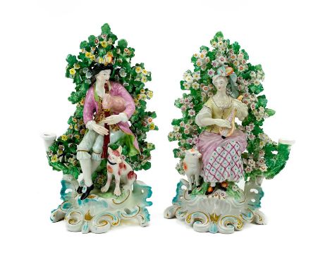 A pair of Derby porcelain bocage candlestick figures. Circa 1780, modelled as musicians, the male seated beside a dog, his fe