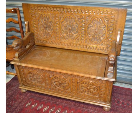 A carved oak monks bench, the moulded edge fold over top on recumbent lion supports, panelled floral decorated front on bun f