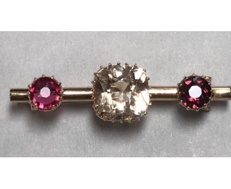 A topaz and ruby three stone bar brooch, the cylindrical bar claw set to the centre with a very pale apricot cushion cut topa