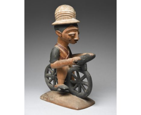 A Yoruba figure riding a bicycle Nigeria wearing a pith helmet and previously with a pipe in his mouth, with painted decorati