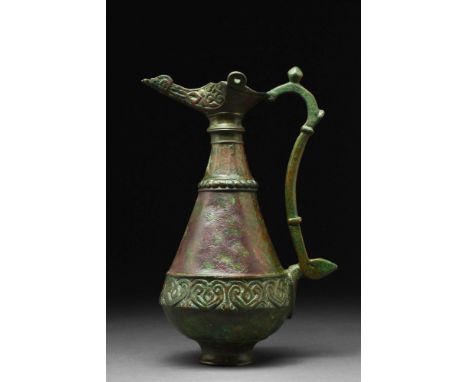 Ca. AD 1100 - 1200. A bronze ewer with an inverted pyriform body, resting on a low foot. It is adorned with a mouth modelled 
