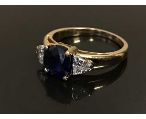 An 18ct gold sapphire and diamond ring, the cushion-cut sapphire within a four claw setting flanked by two trilliant-cut diam