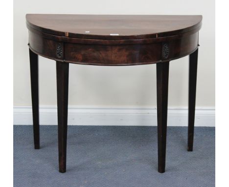 A George III mahogany fold-over demi-lune card table, the frieze carved with oval rosettes, raised on square tapering legs, w