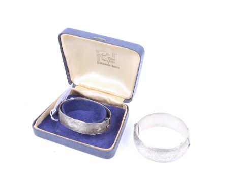 Two silver hinged bangles. Comprising; a shallow flat section bangle part engraved with flowers and trailing leaves, hallmark