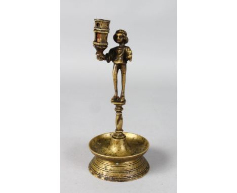 A BRASS CANDLESTICK with a figure holding a candle sconce on a circular base. 8.5ins high.