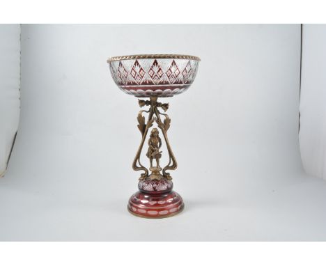 An overlaid ruby glass centre piece, the circular bowl with metal rim supported by a brass figure of a girl with basket, 35cm
